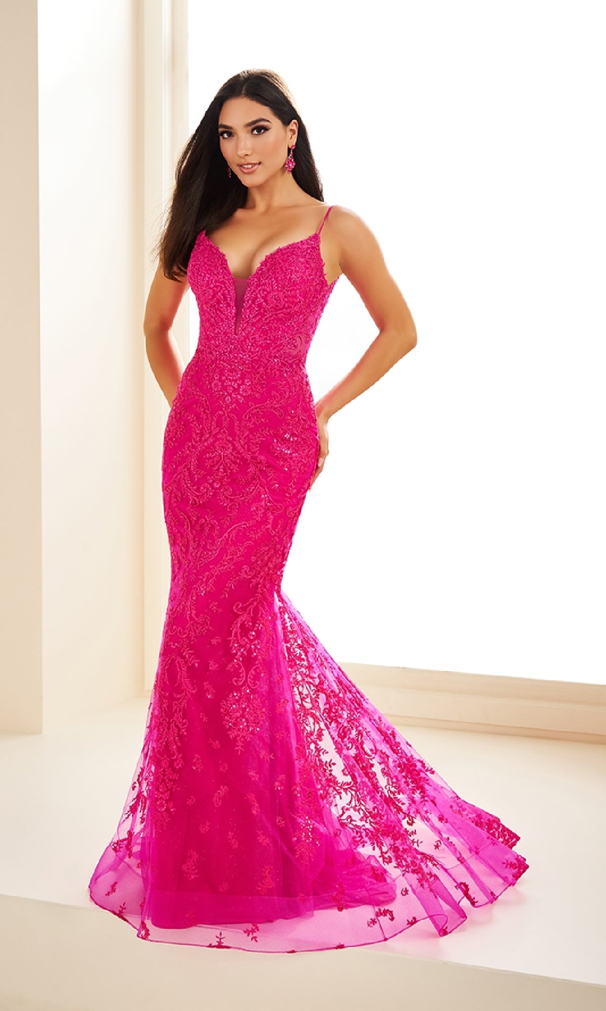 A woman stands indoors wearing the Ellie Wilde EW36096, a bright pink, form-fitting long prom dress with lace embroidery and a sheer flared mermaid hem.